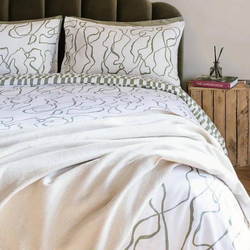 Mantita espiga beige de Morfeo extendida sobre una cama con ropa estampada y cabecero de terciopelo verde.