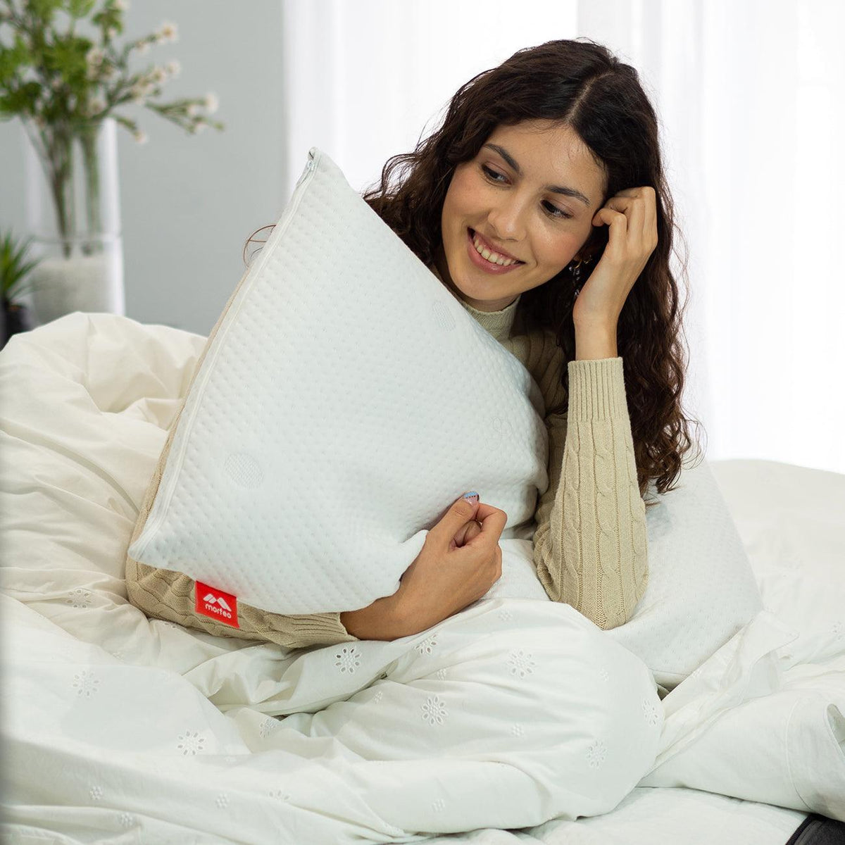 Mujer sonriente abrazando la almohada viscoelástica SojaVisco de Morfeo sentada en la cama.